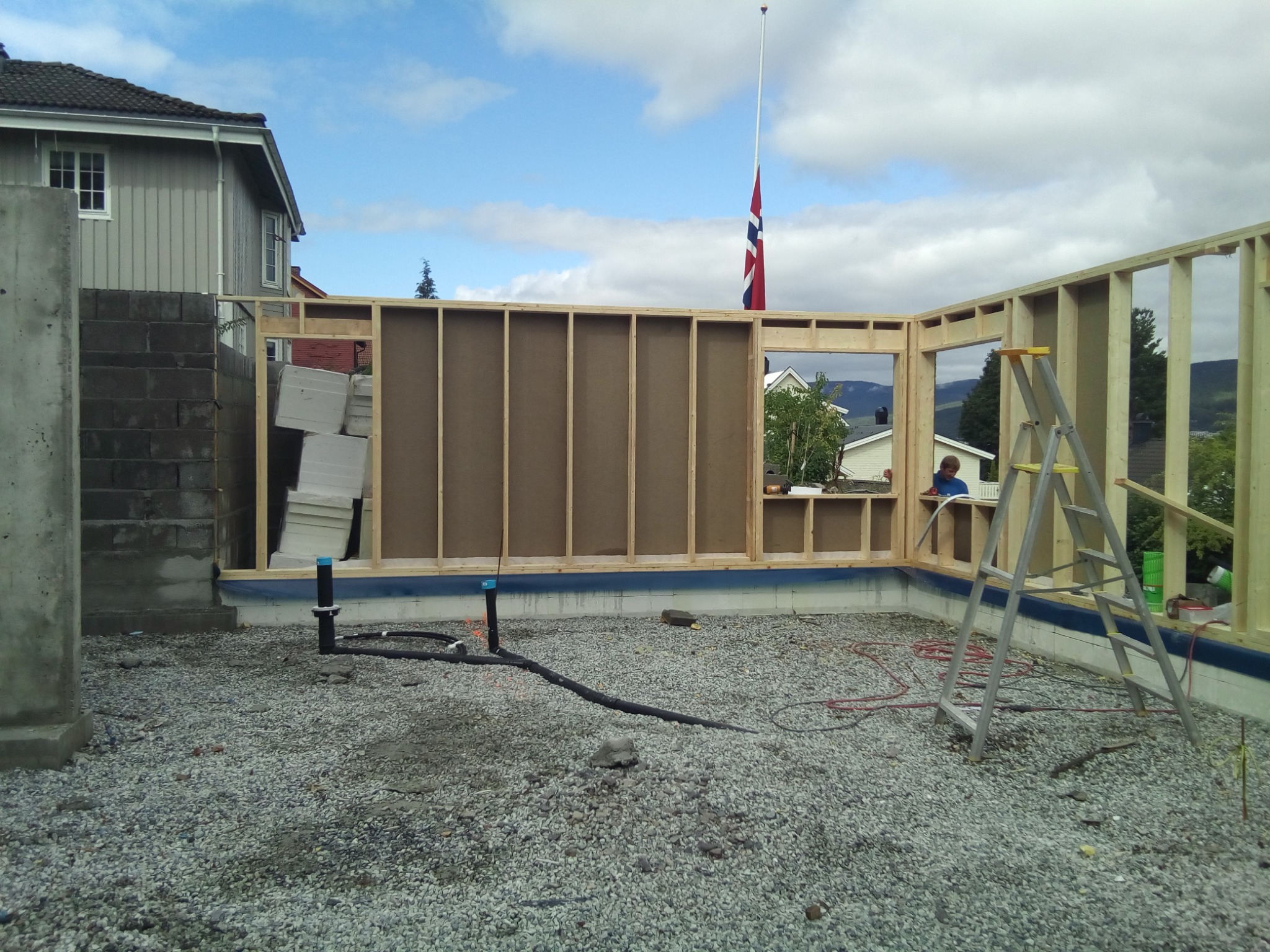 Wznoszenie konstrukcji szkieletowej domu na fundamencie, widoczne elementy konstrukcyjne, instalacje hydrauliczne, izolacja, drabina, w tle budynki mieszkalne i flaga Norwegii na maszcie.