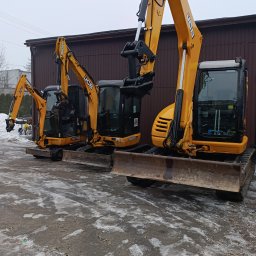 Marcin Dziarnowski - Trzy żółte koparki JCB zaparkowane obok brązowego budynku w pochmurny dzień. Widoczne detale maszyn i otoczenia.