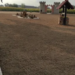 Przygotowany teren pod trawnik z elementami dekoracyjnymi: studnia z daszkiem, miniatura zamku i skalniak z krzewami. W tle rząd tuj.