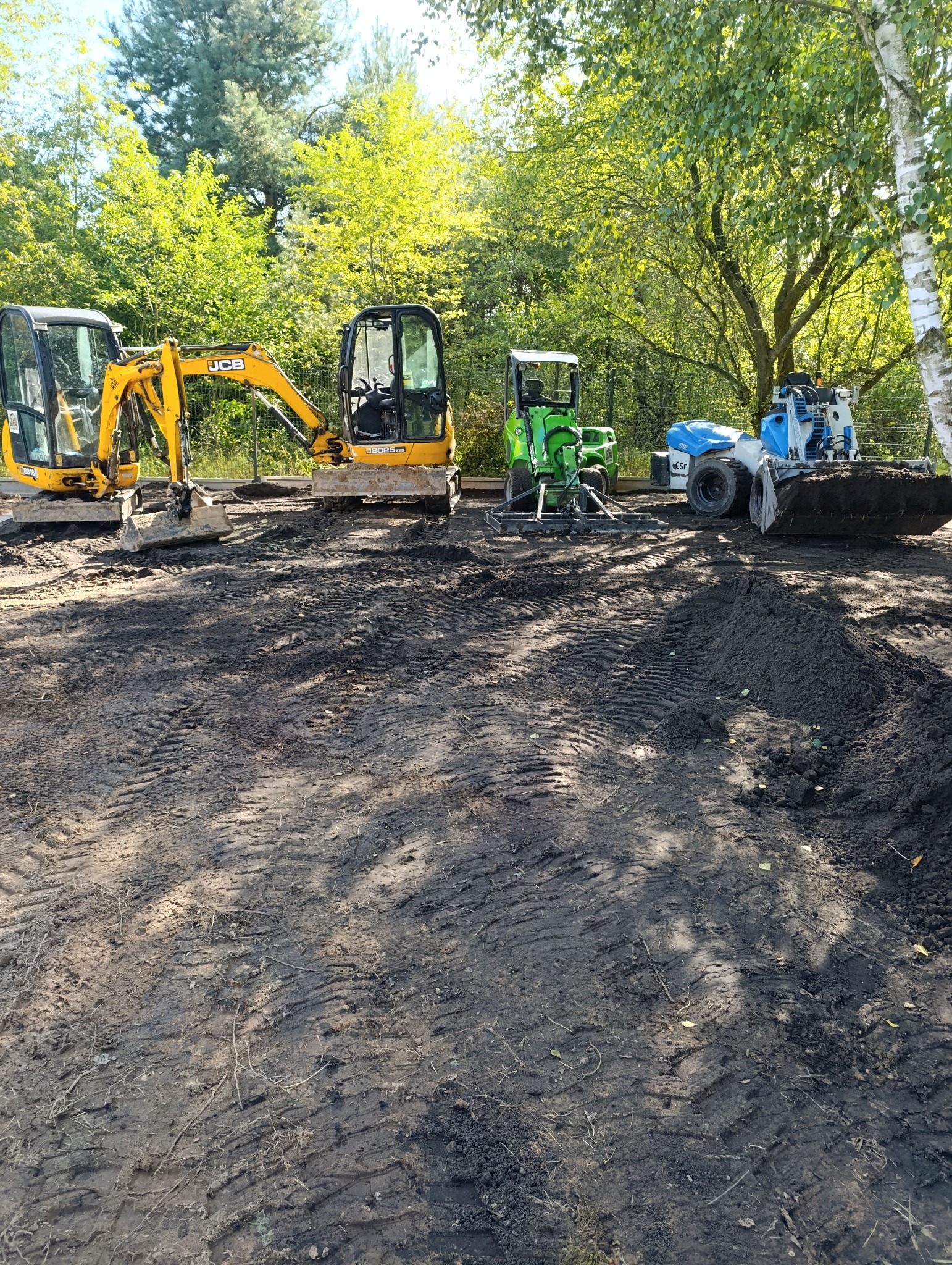 Żółte i zielone mini koparki oraz walec na przygotowanym terenie pod budowę, widoczne ślady opon na ziemi, w tle drzewa.