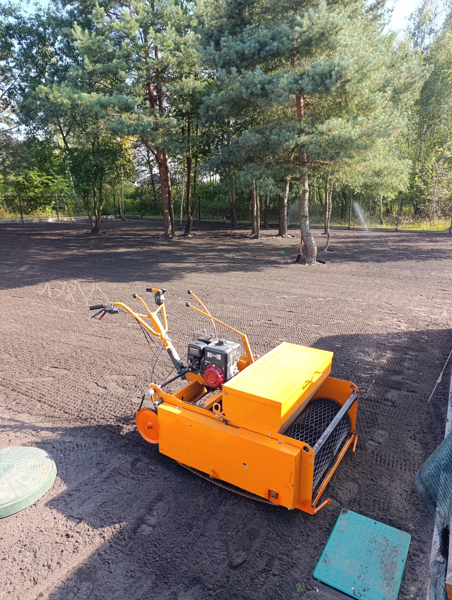 Pomarańczowa maszyna do siewu trawy na przygotowanym terenie. W tle drzewa i system nawadniania w trakcie pracy. Widok z góry na maszynę i teren.