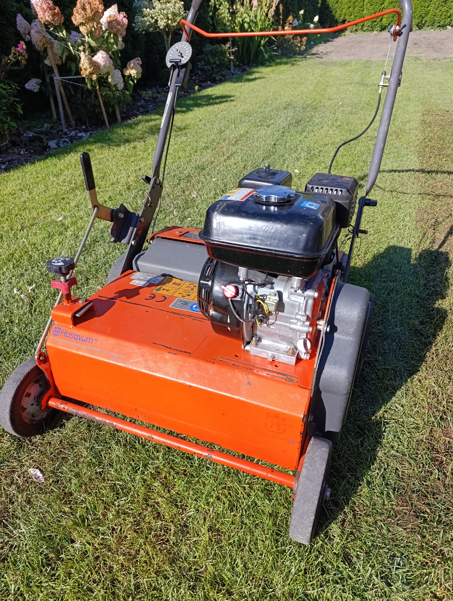 Aerator spalinowy Husqvarna na trawniku. Pomarańczowa maszyna do wertykulacji trawy, widoczne detale silnika i kół. W tle kwiaty hortensji.