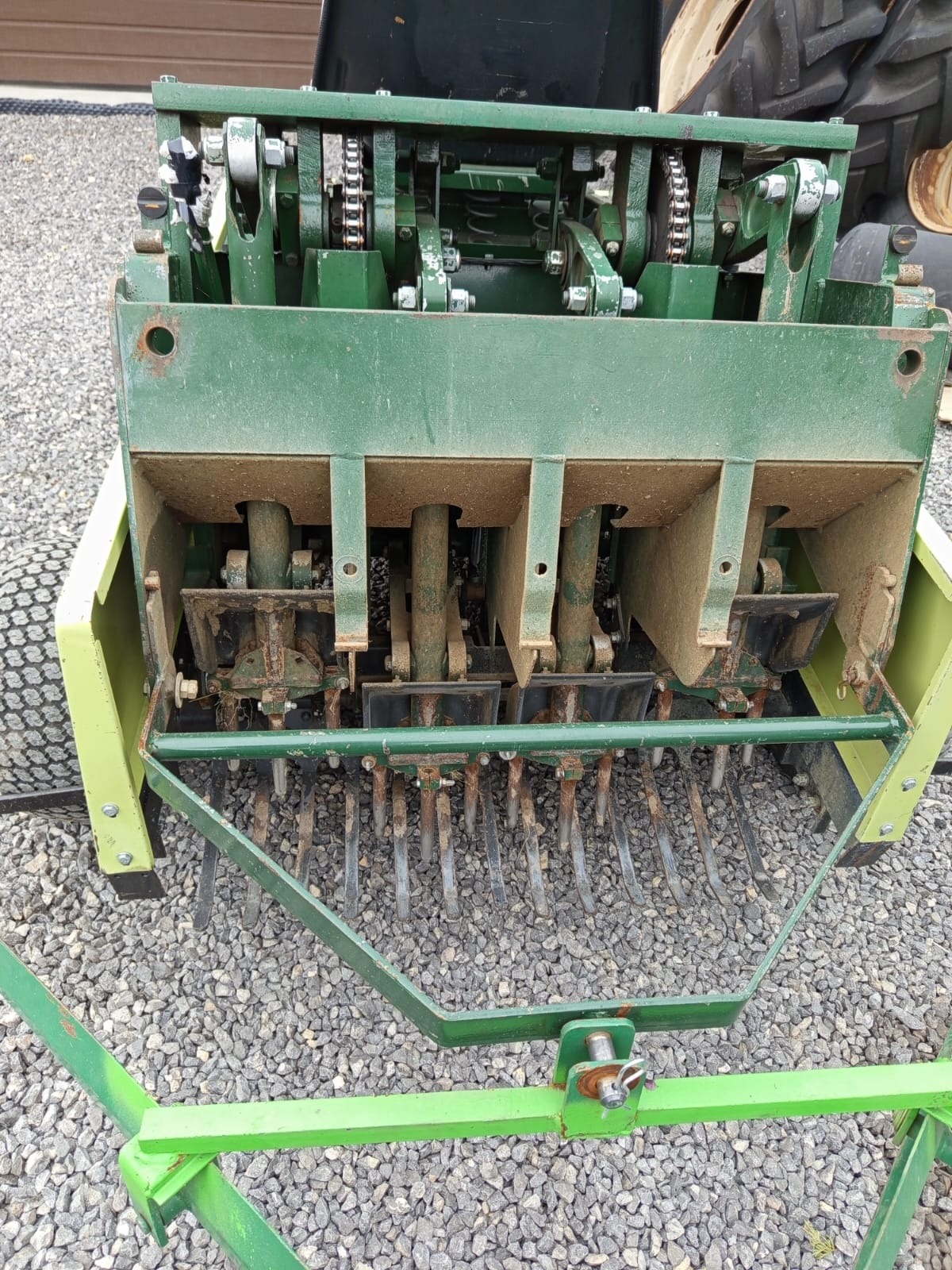 Aerator do trawy z bliska, z widocznymi ostrzami i mechanizmem łańcuchowym, na tle żwirowego podłoża. Sprzęt w odcieniach zieleni, z elementami rdzy.