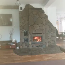 Kamienny kominek z paleniskiem w salonie, drewniana podłoga i dywan, dekoracyjne lampy wiszące.