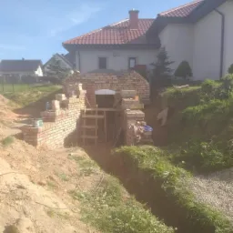 Budowa piwnicy z cegły z łukowym wejściem, widoczny fragment domu w tle, teren w trakcie prac ziemnych z wykopem i roślinnością.