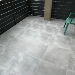 Szare płytki podłogowe na zewnątrz, na tarasie, z ciemnym, poziomym ogrodzeniem i zielonym krzesłem w rogu.