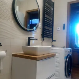 Nowoczesna łazienka z owalnym lustrem z podświetleniem LED, umywalką nablatową na szafce z drewnianym blatem, czarną armaturą i grzejnikiem drabinkowym, obok pralka.