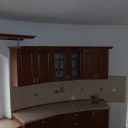 Klasyczne, ciemne drewniane szafki kuchenne z witrynami, beżowe płytki nad blatem z owocowymi motywami, zaokrąglony blat, widok na okno z widokiem na zewnątrz, dekoracyjny gzyms sufitowy.