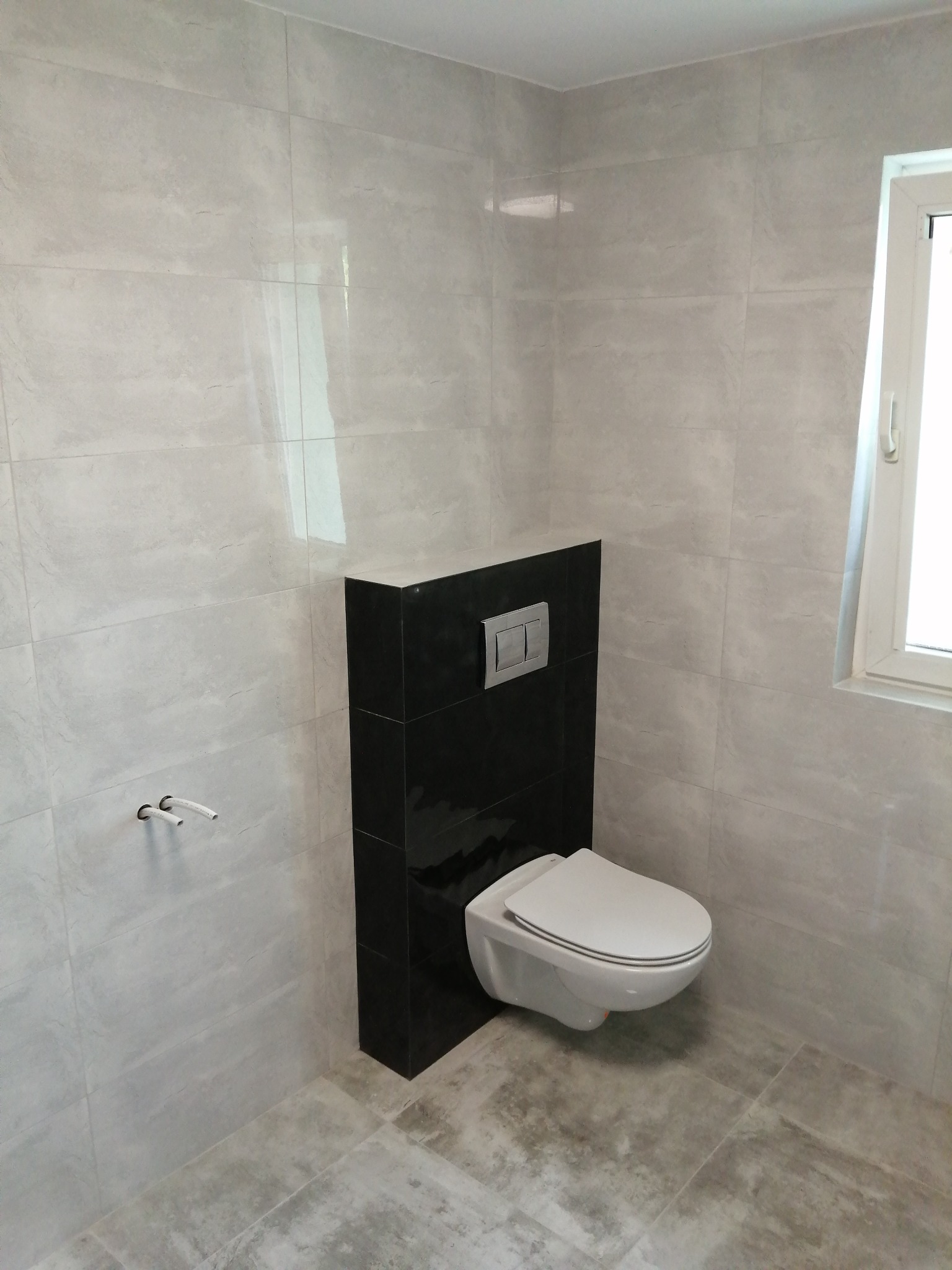 Nowoczesna łazienka z podwieszaną toaletą na czarnym stelażu, szare płytki na ścianach i podłodze, widoczne przyłącza hydrauliczne i okno.