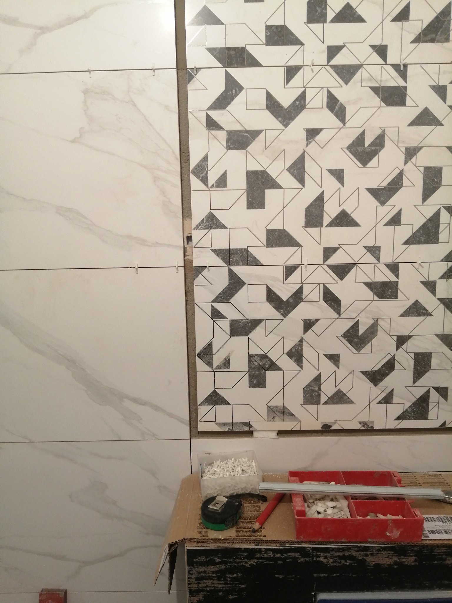 Układanie płytek: połączenie białych płytek imitujących marmur z dekoracyjnymi płytkami w geometryczne wzory, widoczne krzyżyki dystansowe i narzędzia na kartonie.