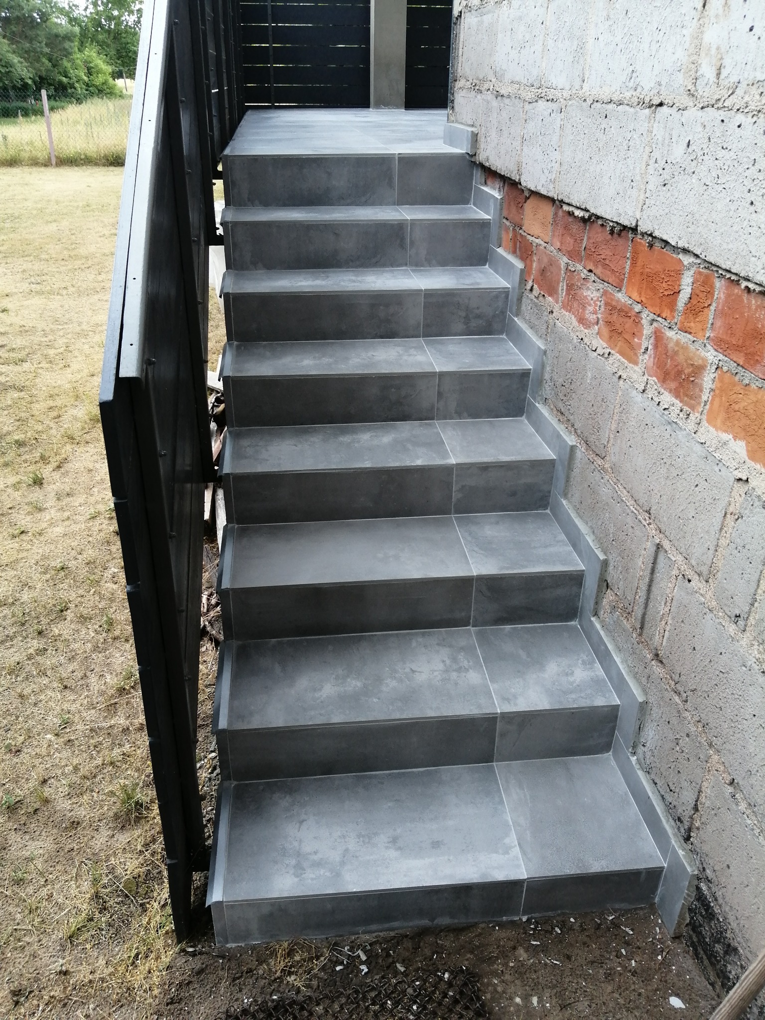 Zewnętrzne schody wyłożone szarą, kwadratową glazurą, z czarną metalową barierką po lewej stronie i murem z cegieł po prawej, prowadzące do drzwi.