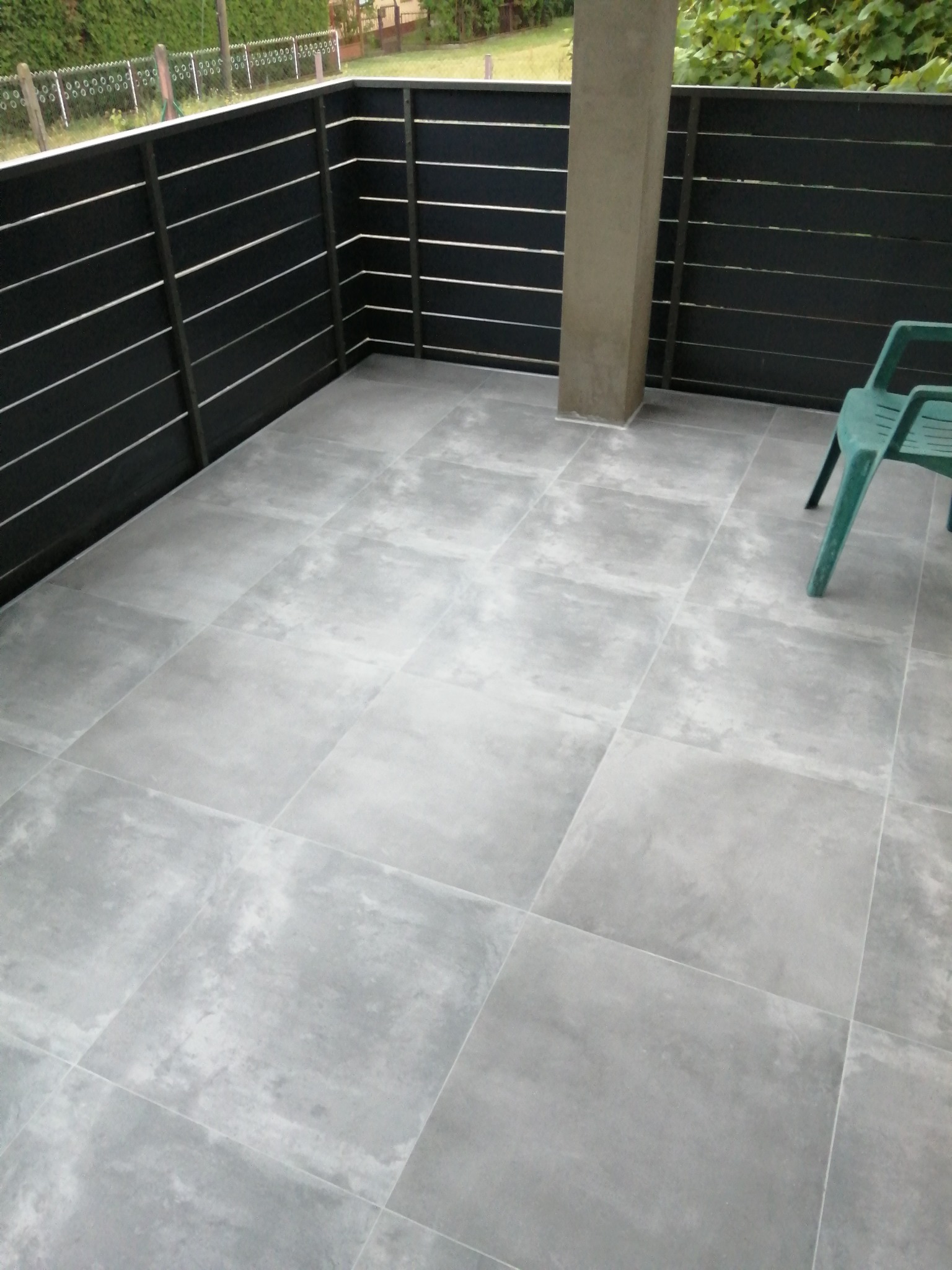 Szare płytki podłogowe na zewnątrz, na tarasie, z ciemnym, poziomym ogrodzeniem i zielonym krzesłem w rogu.