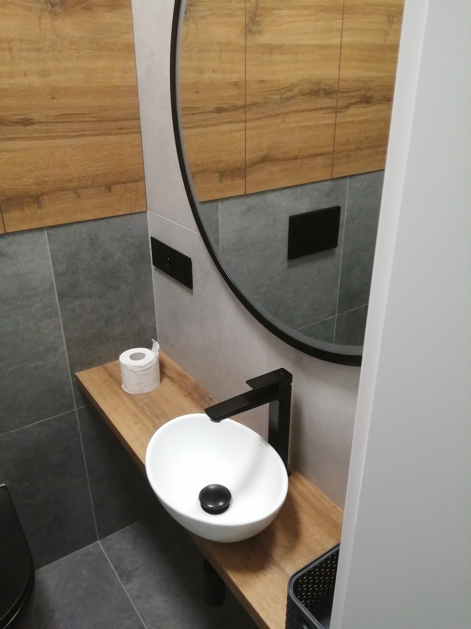 Nowoczesna łazienka z umywalką nablatową, czarną armaturą i okrągłym lustrem, drewnopodobne szafki wiszące, szare płytki na ścianach i podłodze, rolka papieru toaletowego na półce.