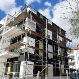 Pracownik na rusztowaniu podczas wykonywania elewacji budynku z panelami w kolorach białym, czarnym i żółtym, widok z dołu na błękitne niebo z chmurami.