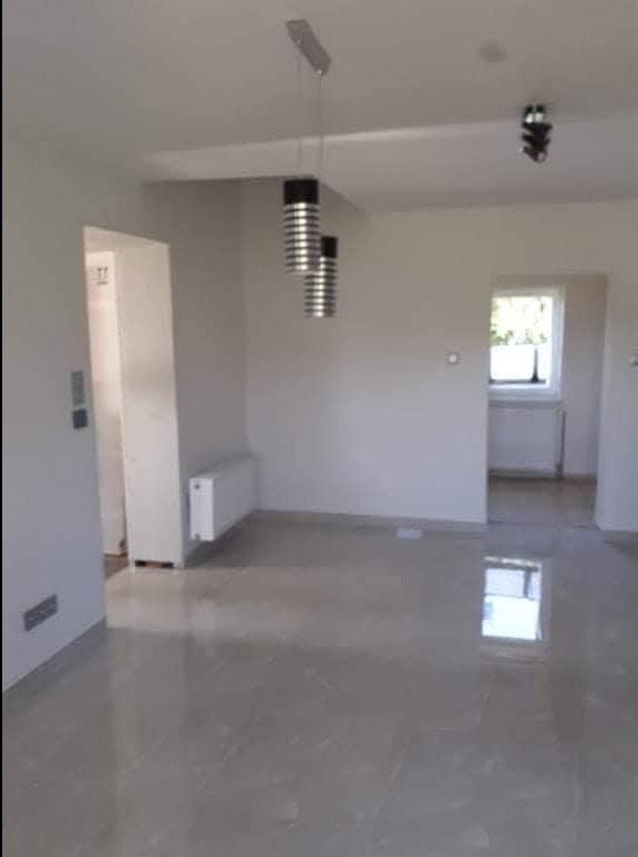 Jasne wnętrze po remoncie: błyszcząca podłoga z płytek, białe ściany, nowoczesne lampy wiszące i grzejnik. Widok na sąsiednie pomieszczenie przez otwarte drzwi.