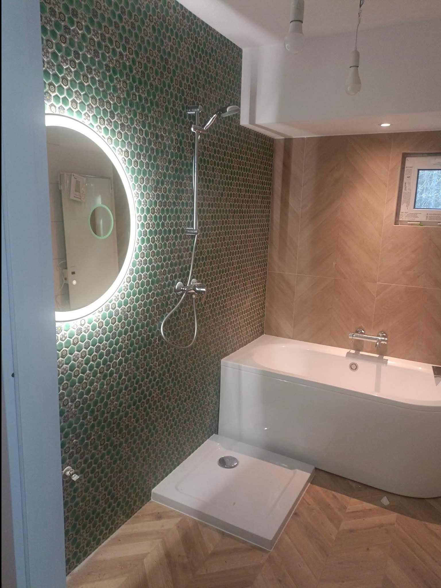 Nowoczesna łazienka z zieloną mozaiką heksagonalną, okrągłym lustrem z podświetleniem LED, białą wanną i brodzikiem. Podłoga wyłożona panelami w jodełkę.