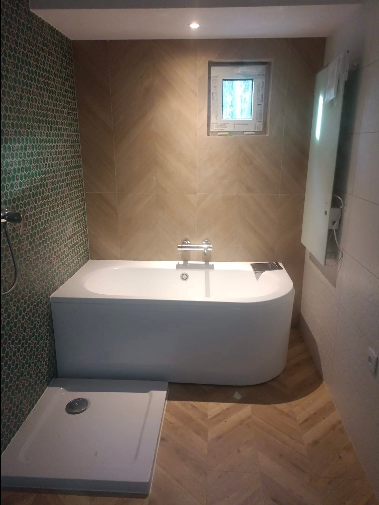 Nowoczesna łazienka po remoncie: biała wanna, brodzik, mozaika na ścianie i płytki imitujące drewno ułożone w jodełkę. Okno w tle.