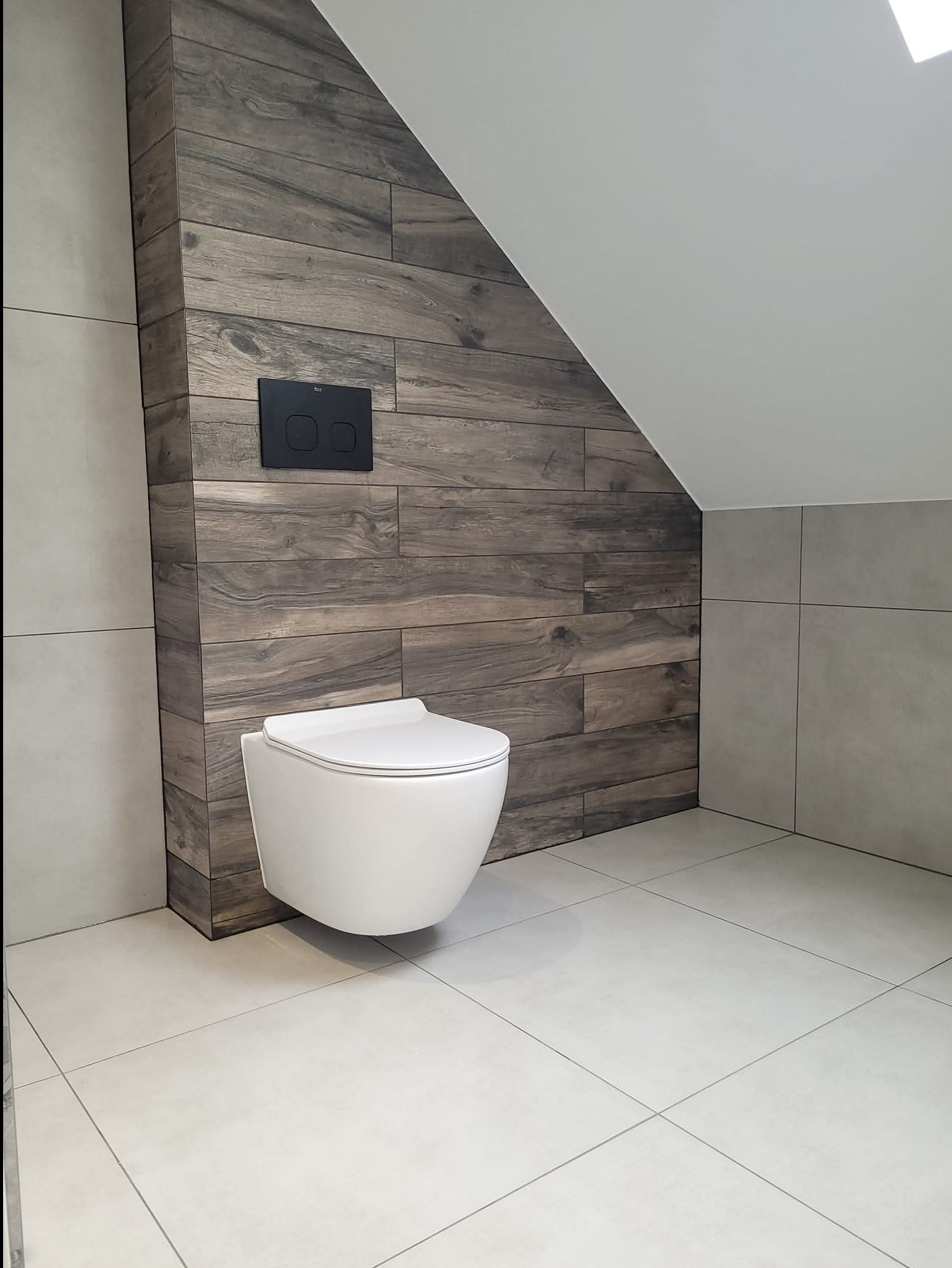 Nowoczesna łazienka na poddaszu: podwieszana toaleta z czarnym przyciskiem spłukującym na tle ściany z płytek imitujących drewno, jasne płytki podłogowe i ścienne.