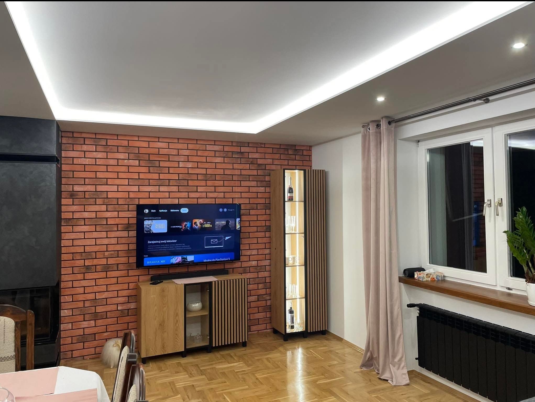 Salon po remoncie: ceglana ściana z telewizorem, drewniana komoda RTV, wysoka witryna z oświetleniem LED, parkiet i podwieszany sufit z listwą LED.