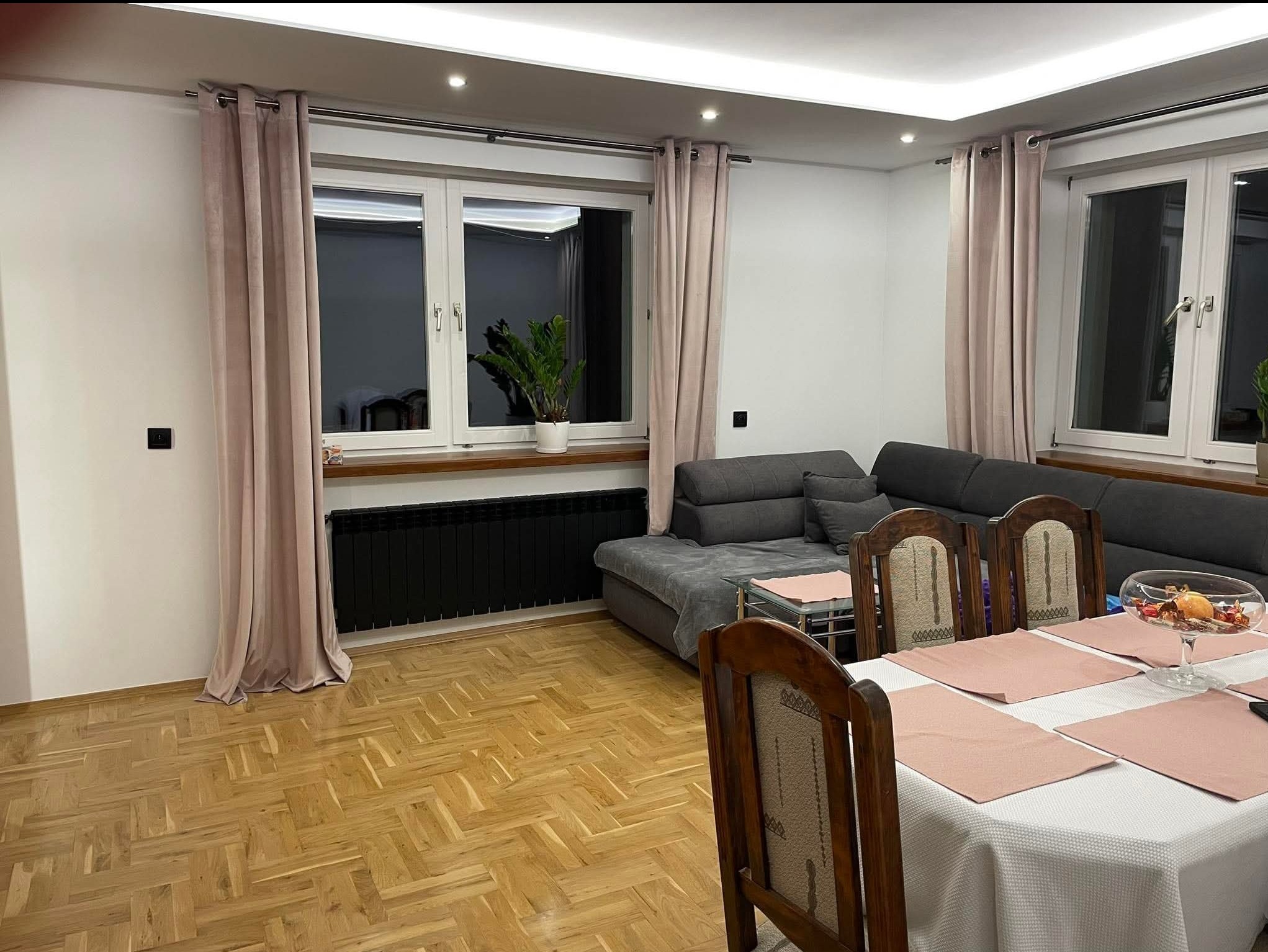 Jasny parkiet w jodełkę, szary narożnik i pudrowo-różowe zasłony w przestronnym salonie z oknami. Na stole dekoracja w szklanym naczyniu.