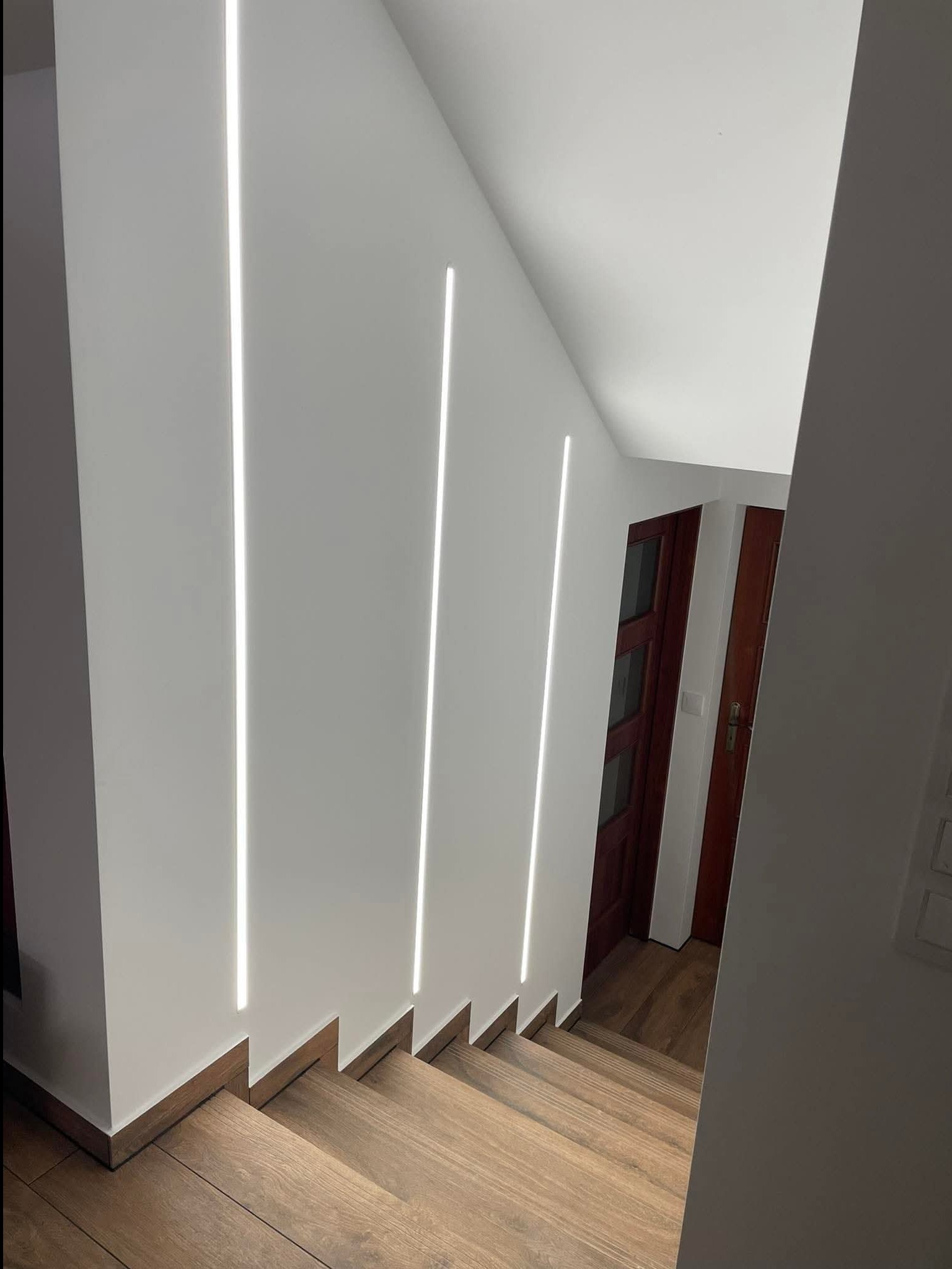 Nowoczesne oświetlenie schodów z trzema pionowymi pasami LED w ścianie, drewniane stopnie i widoczne drzwi w tle. Minimalistyczny design.