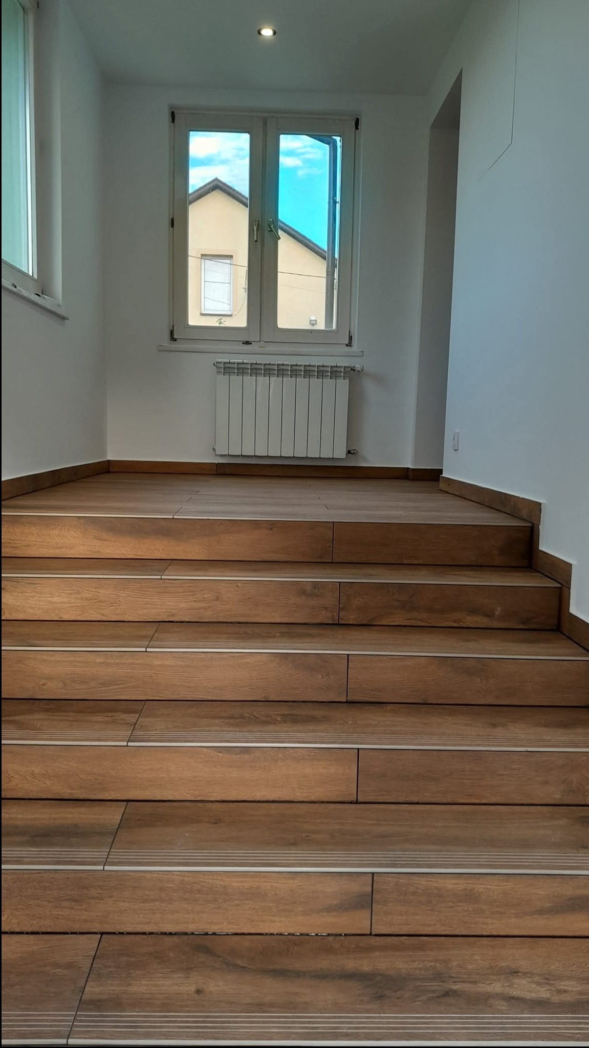 Schody wyłożone płytkami imitującymi drewno, z metalowymi listwami antypoślizgowymi. W tle okno z widokiem na budynek i grzejnik. Minimalistyczne wnętrze.