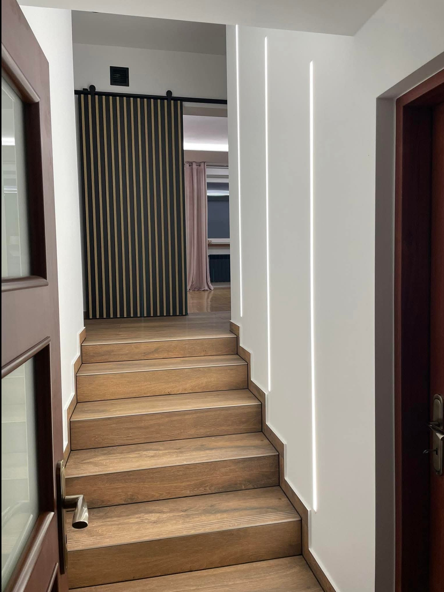 Nowoczesne schody z drewnopodobnymi płytkami prowadzą do wnętrza z przesuwnymi drzwiami lamelowymi i oświetleniem LED wzdłuż ściany.