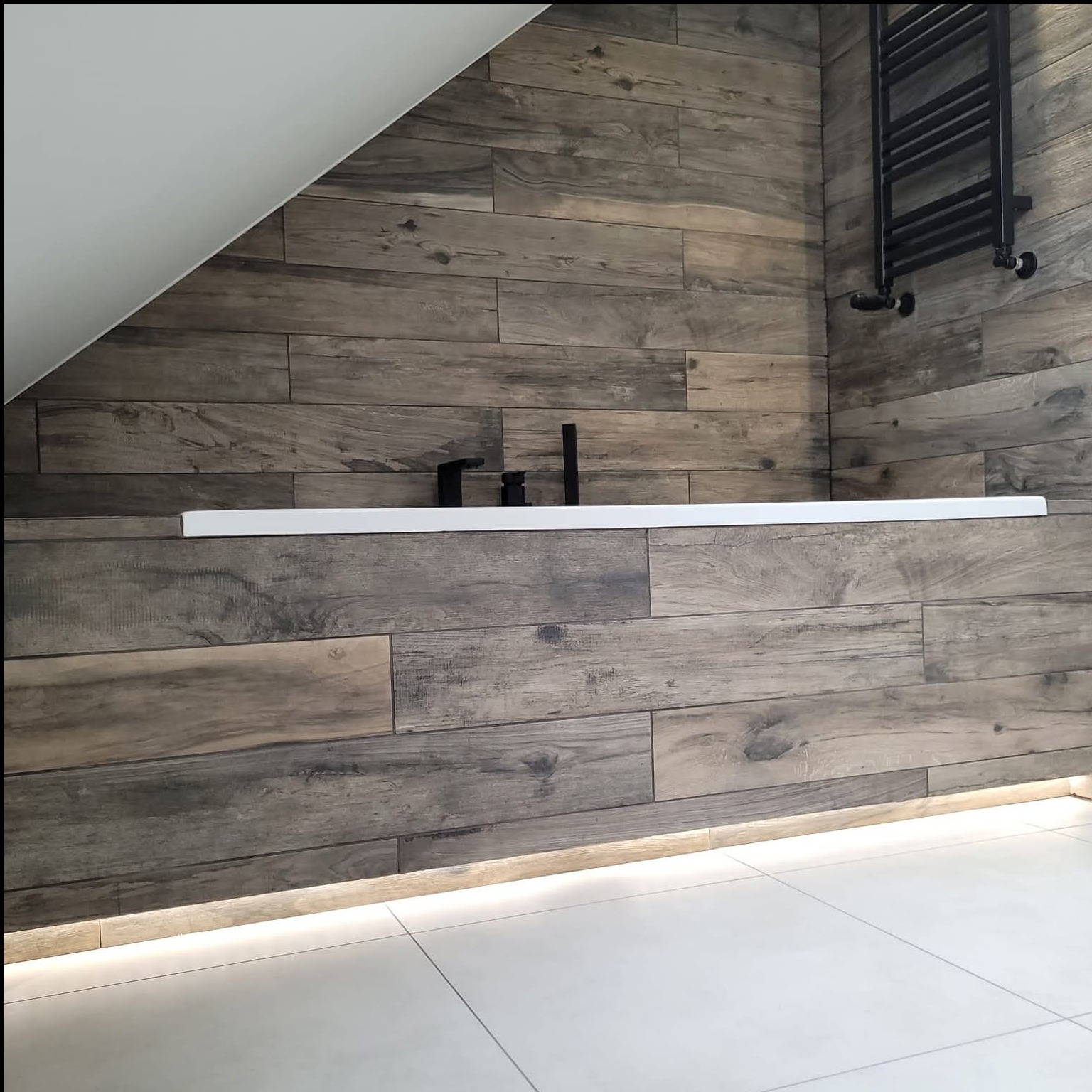 Nowoczesna łazienka na poddaszu z wanną obudowaną płytkami imitującymi drewno, podświetleniem LED i czarnym grzejnikiem drabinkowym.