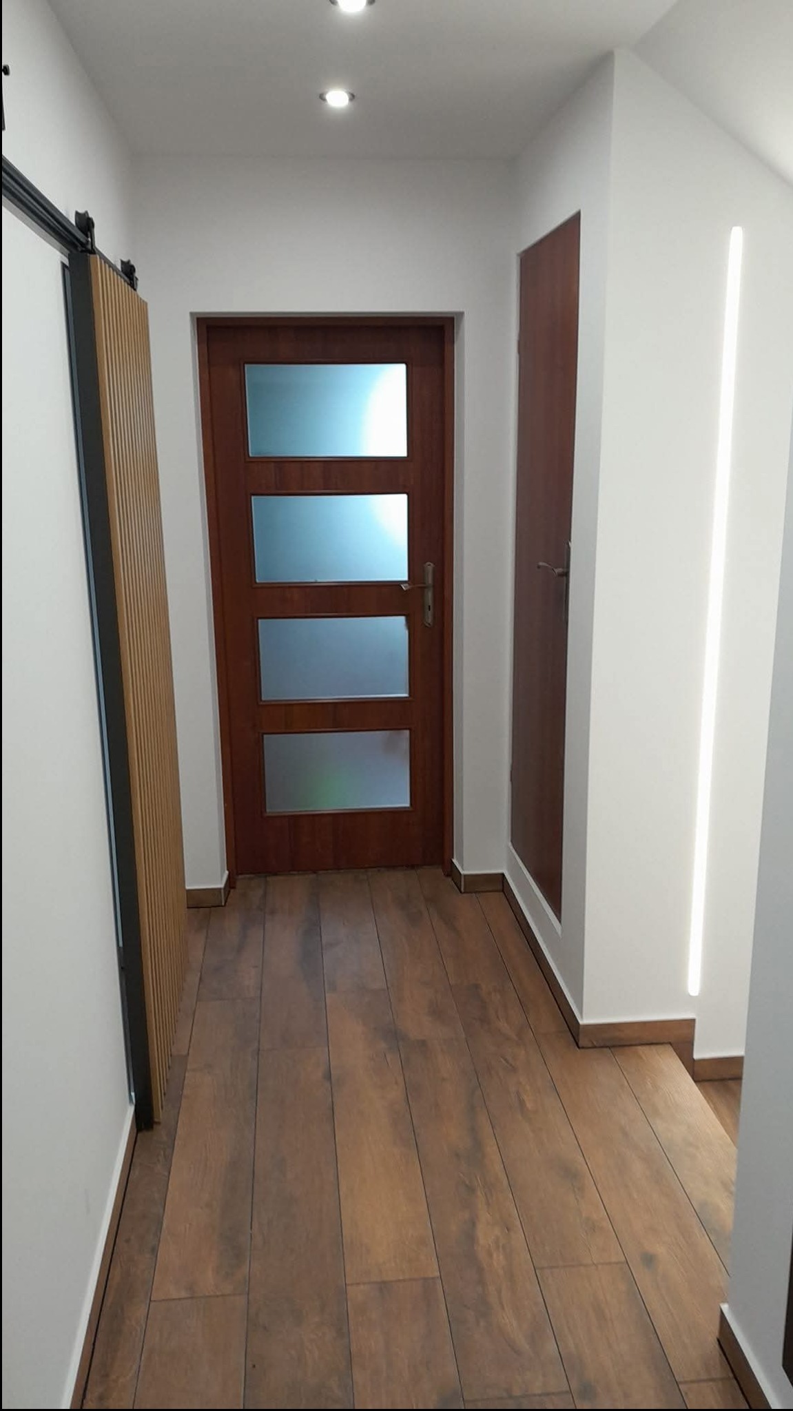 Nowoczesny korytarz z drewnianą podłogą, drzwiami z przeszkleniami i oświetleniem LED. Minimalistyczny design z akcentami drewna i bieli.