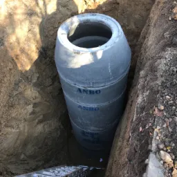 Betonowy zbiornik z napisem ANBO umieszczony w głębokim wykopie, widoczna drabina ułatwiająca zejście na dół.