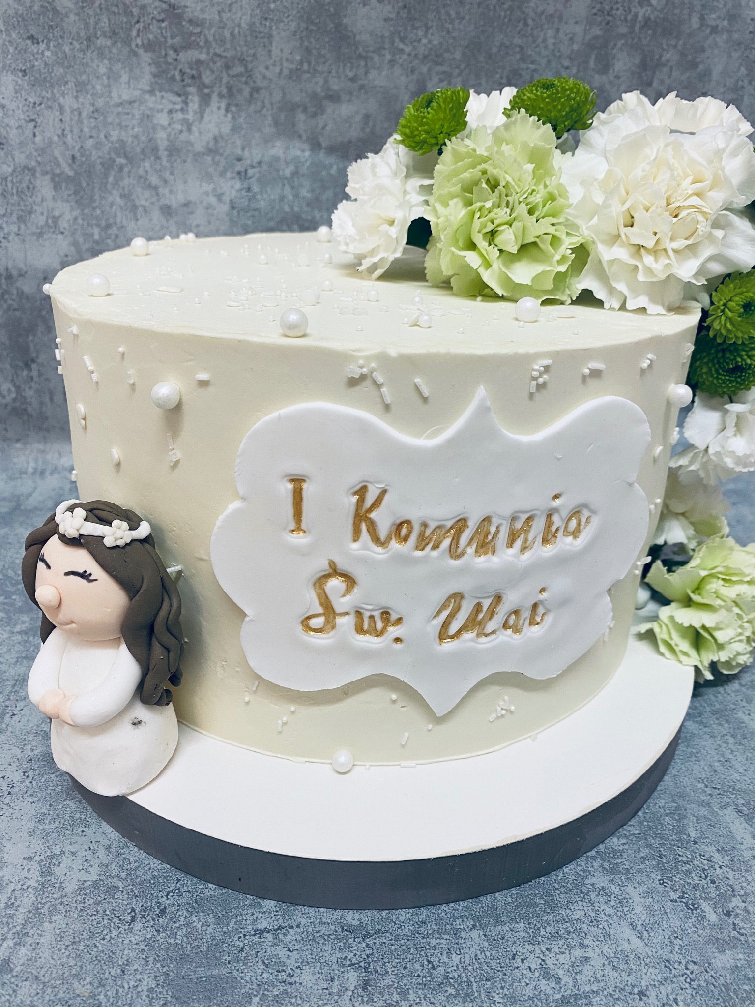 Tort komunijny z białą polewą, dekorowany białymi i zielonymi kwiatami, perłowymi koralikami, oraz figurką dziewczynki w stroju komunijnym i napisem 'I Komunia Święta' w złotym kolorze.