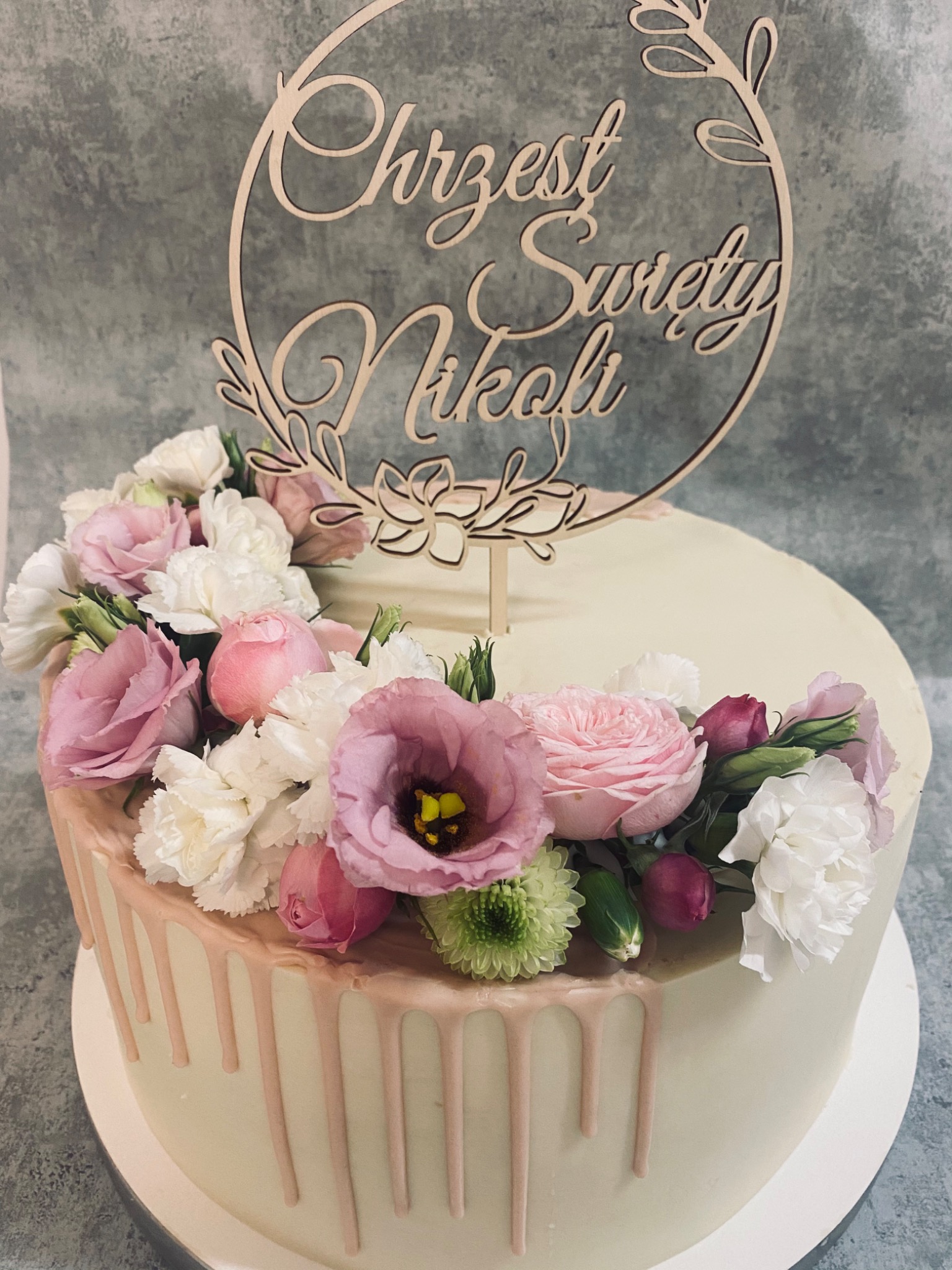 Tort z kremową polewą i dekoracją z żywych kwiatów w odcieniach różu i bieli, zwieńczony drewnianym topperem z napisem 'Chrzest Święty Nikoli'.