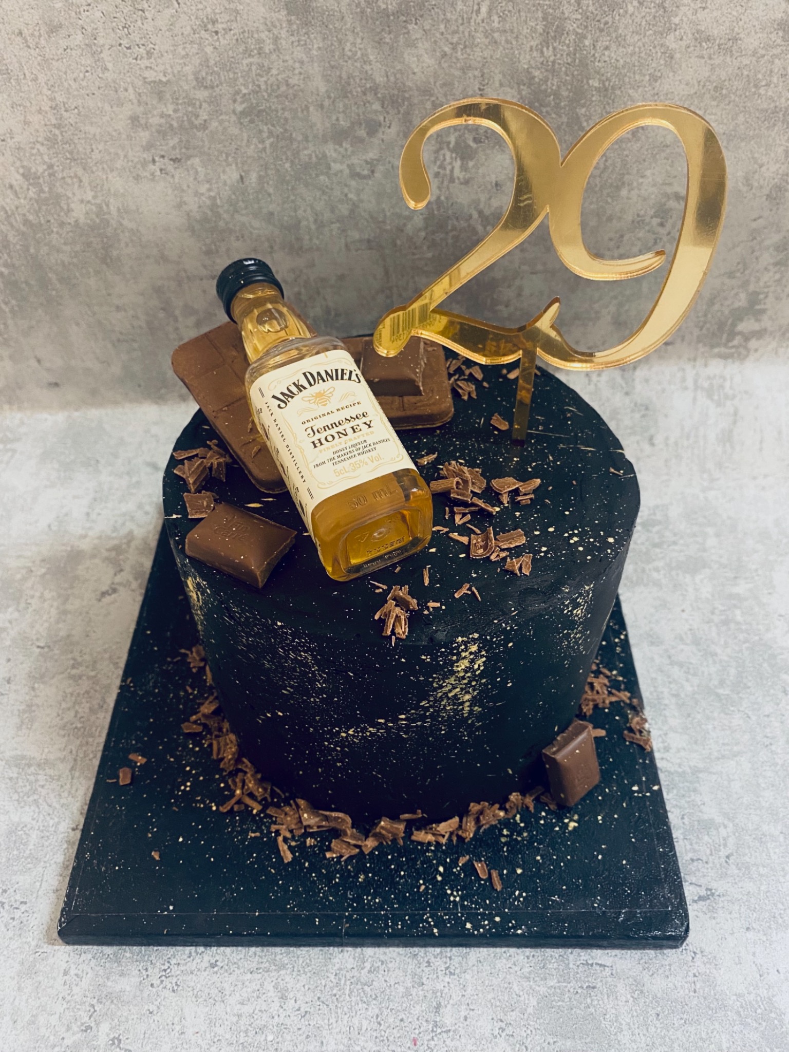 Tort urodzinowy z czarnym lukrem, dekorowany czekoladą, złotym topperem z liczbą 29 i miniaturową butelką Jack Daniel's Tennessee Honey.