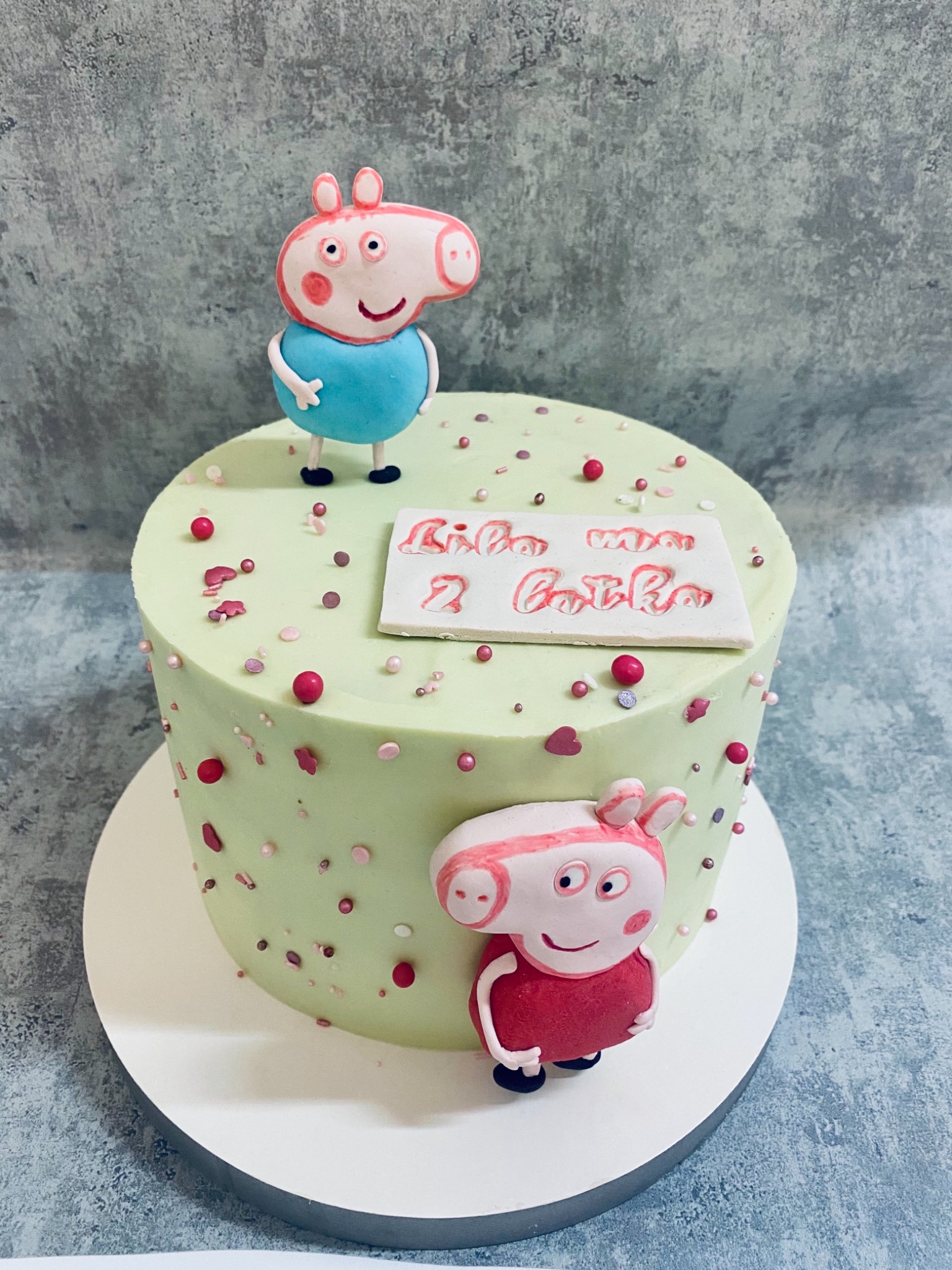 Tort urodzinowy z zielonym lukrem, dekorowany figurkami Peppy i George'a, posypany kolorowymi kuleczkami, z napisem 'Sto lat Zuziu 2 latka' na białej tabliczce.