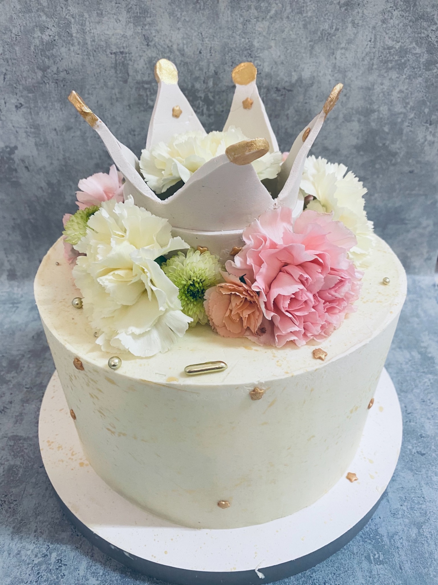 Tort dwuwarstwowy z białą koroną dekoracyjną i kwiatami w odcieniach bieli, różu i zieleni, dodatkowo ozdobiony złotymi elementami i perłami.