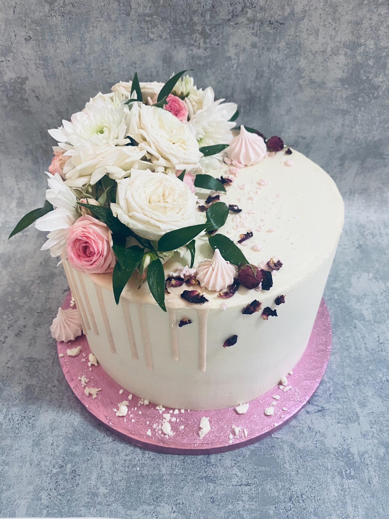 Tort z białym kremem, udekorowany kompozycją z białych i różowych kwiatów, różowych bezików i suszonych płatków róż, z delikatnymi zaciekami z kremu na bokach, umieszczony na różowej podkładce.
