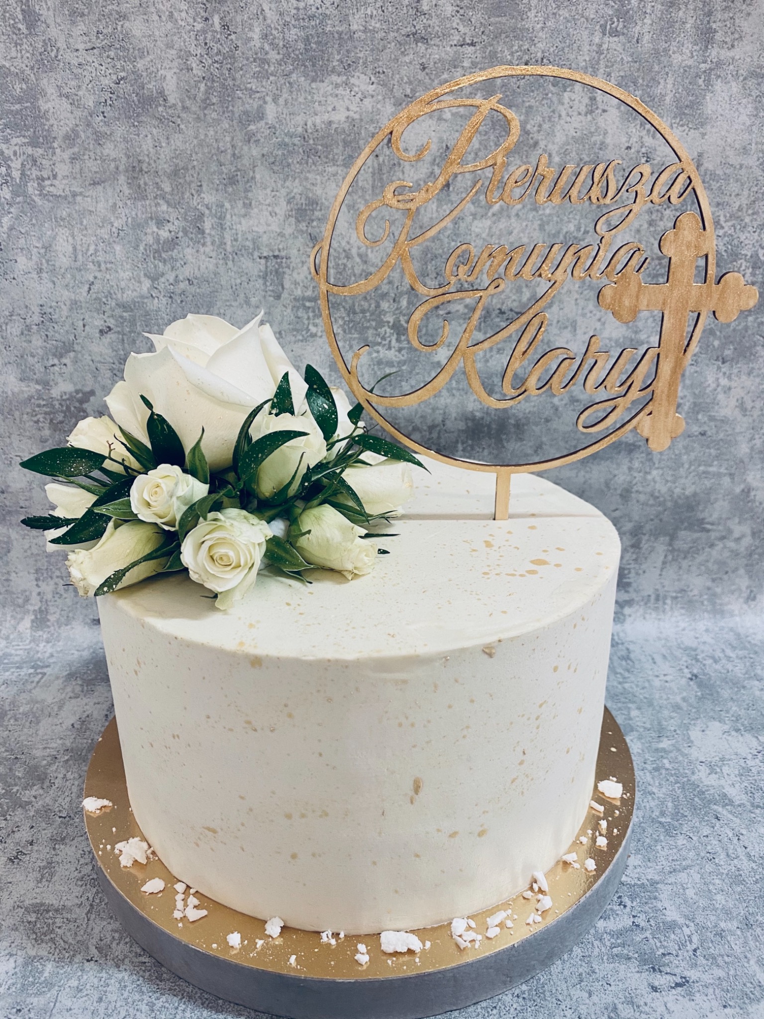 Tort komunijny z białym kremem, dekorowany bukietem z białych róż i złotym topperem z napisem i krzyżem.