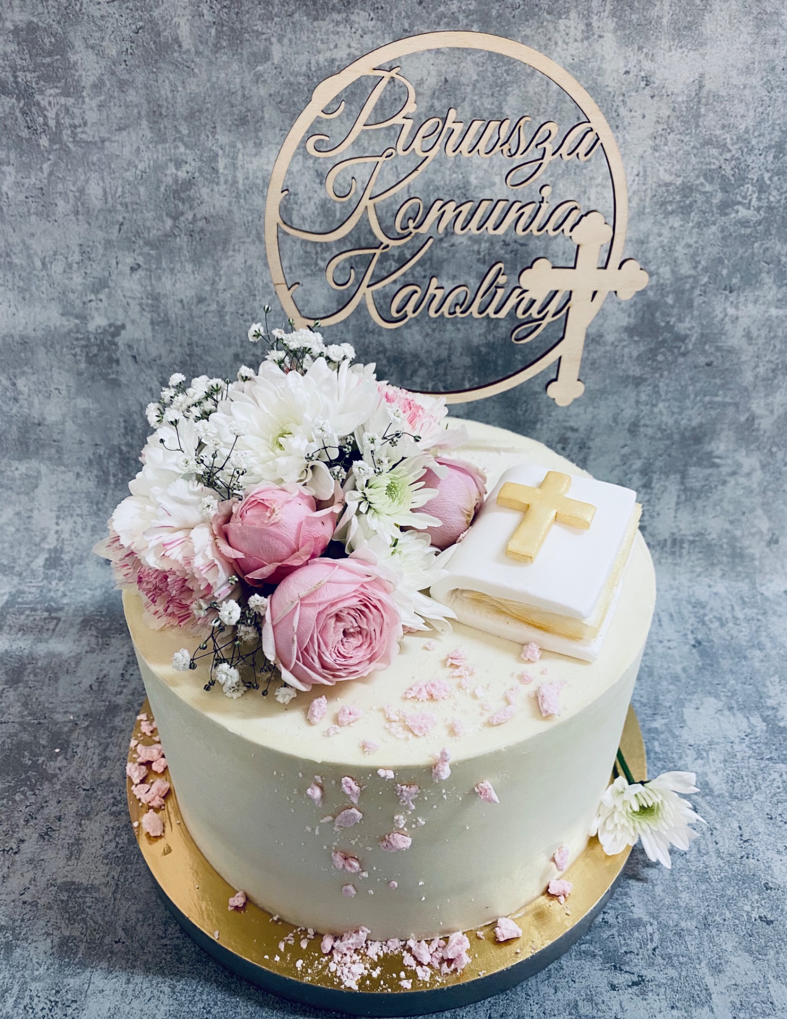 Tort komunijny z białym kremem, dekorowany różowymi różami, białymi chryzantemami i drewnianym topperem z napisem 'Pierwsza Komunia Karoliny' oraz marcepanową książeczką z krzyżem.