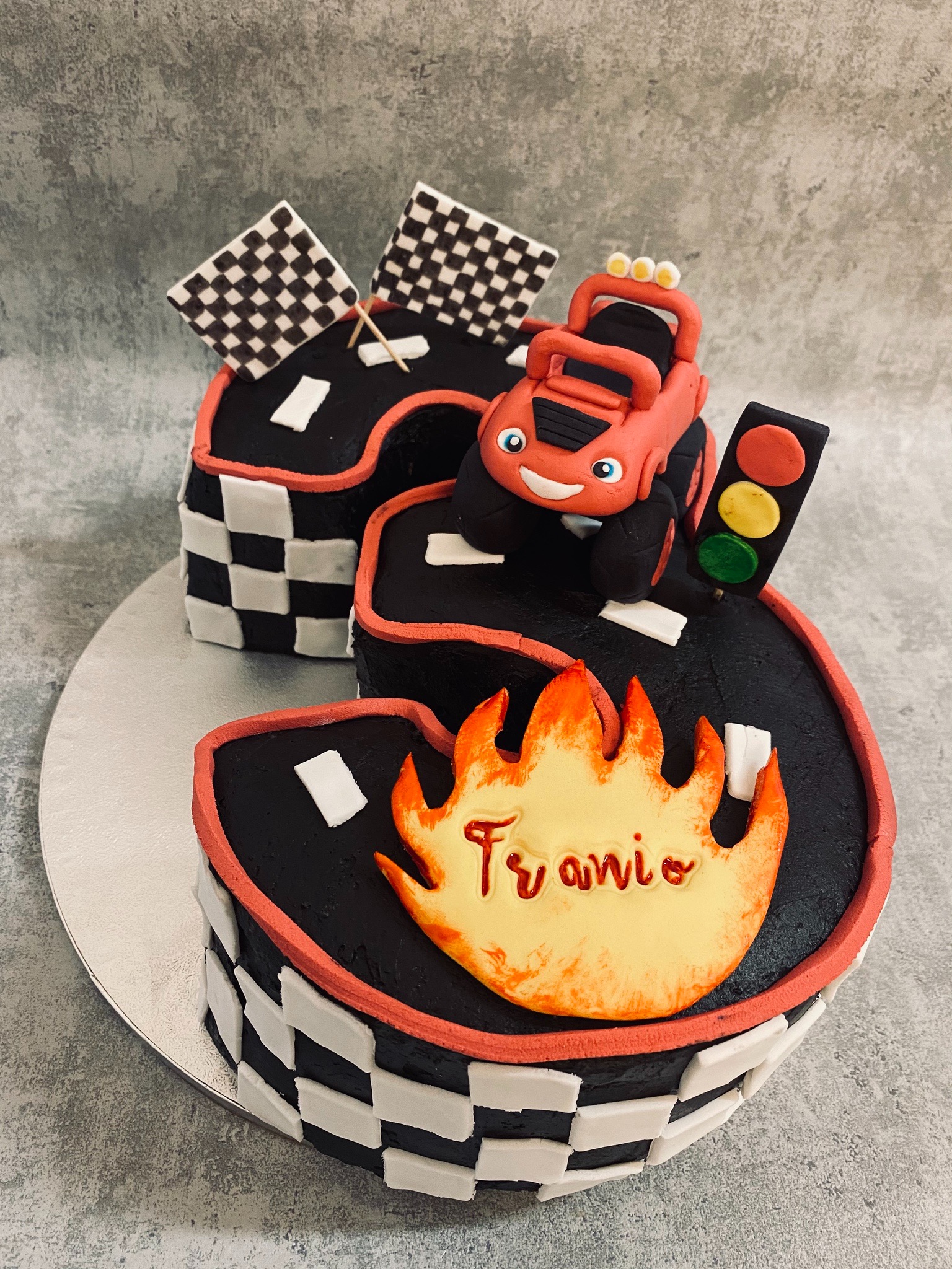 Urodzinowy tort w kształcie toru wyścigowego z dekoracją w postaci płomienia z napisem 'Franio', czerwonej terenówki z uśmiechem i sygnalizacji świetlnej.