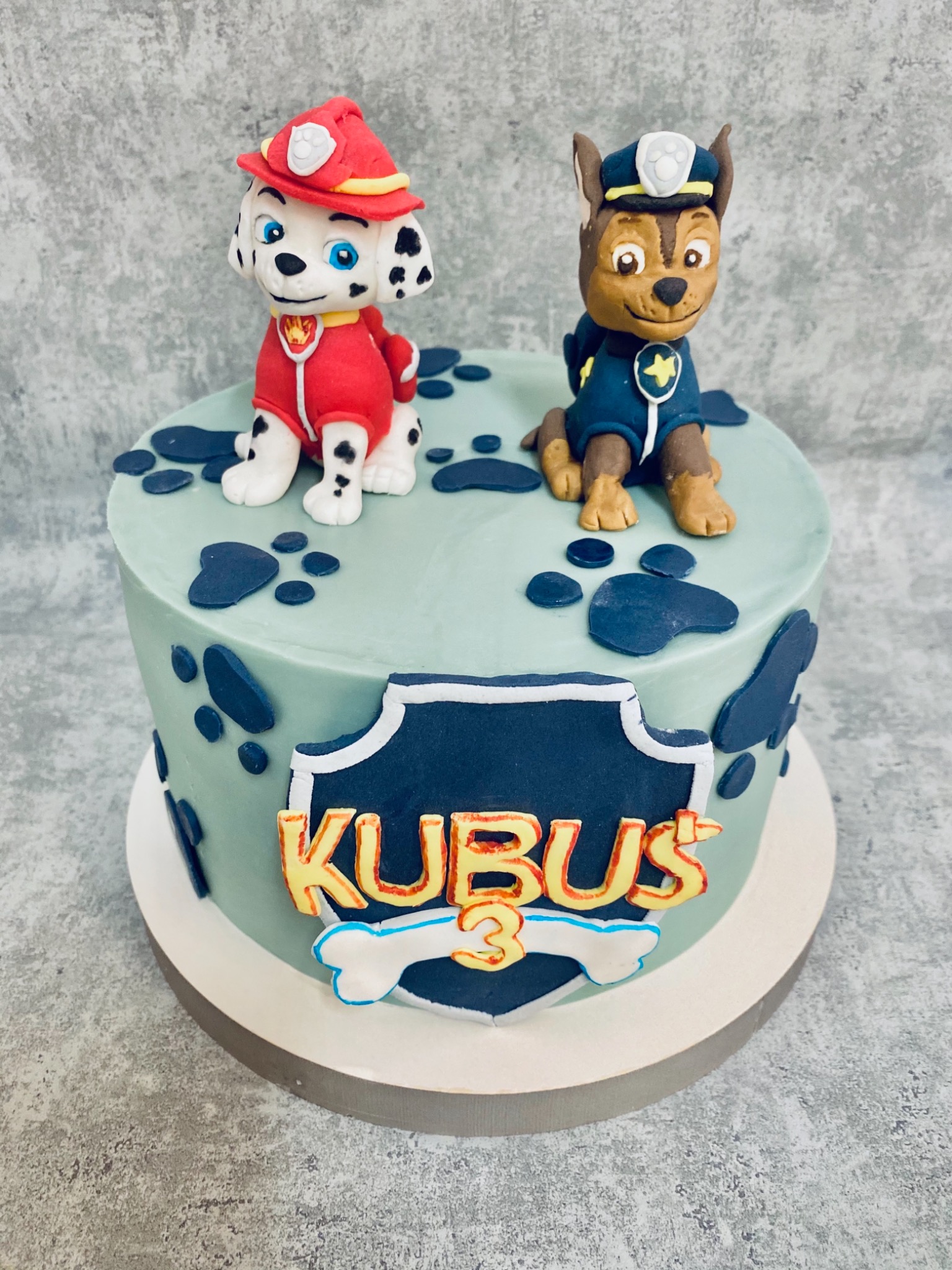 Tort urodzinowy dla dziecka z figurkami Marshalla i Chase z Psiego Patrolu, imię 'KUBUS' i cyfra '3' na ciemnoniebieskim tle w kształcie tarczy, dekorowany ciemnoniebieskimi odciskami łap...