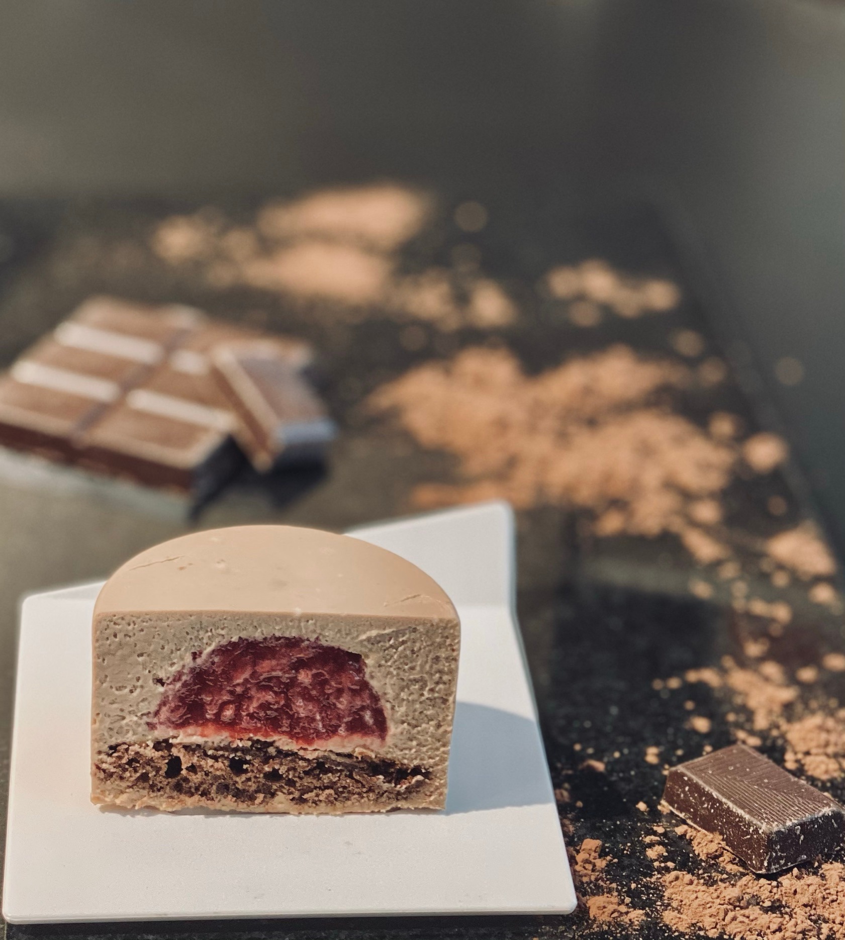 Przekrój monoporcji czekoladowej z musem truskawkowym na białym talerzyku, obok kostka czekolady i rozsypane kakao na ciemnym blacie.