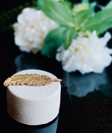 Elegancki, mały torcik z białym, aksamitnym wykończeniem, ozdobiony złotym, jadalnym piórkiem na tle białych kwiatów i ciemnej, błyszczącej powierzchni.