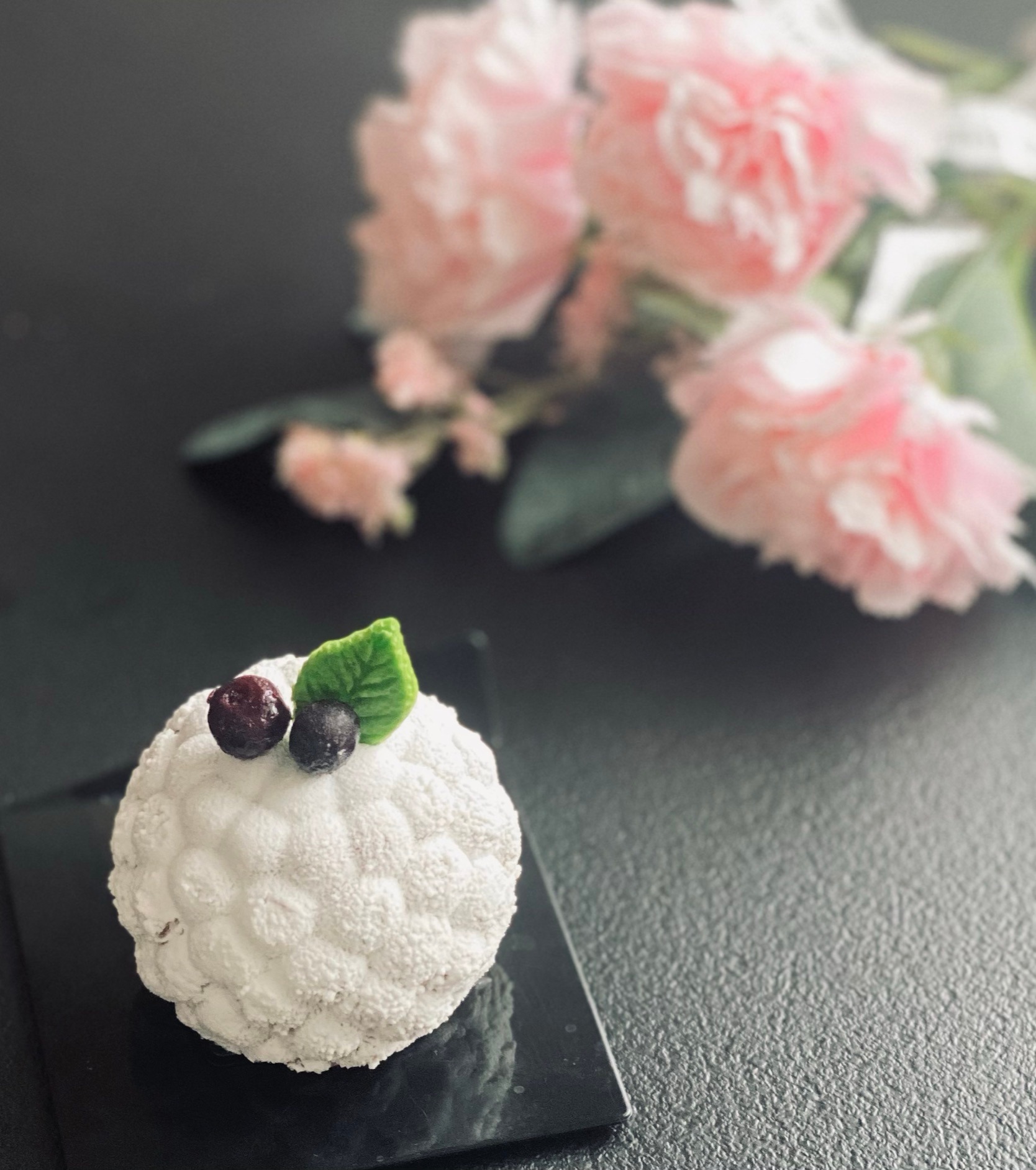 Białe, kuliste ciastko z owocami i listkiem na czarnym talerzyku, w tle rozmyte różowe kwiaty.
