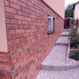 Elewacja budynku wykończona czerwoną cegłą klinkierową, widoczny fragment okna i chodnik z kostki brukowej prowadzący w górę, obok rosną krzewy róż.