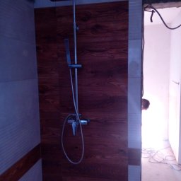 Firma budowlana MC-POL Marian Cielecki - Kabina prysznicowa z panelem imitującym drewno, chromowana bateria z deszczownicą, widoczne przewody elektryczne i fragment osoby w przejściu do innego pomieszczenia.