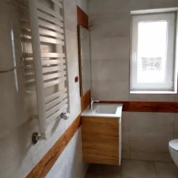 Nowoczesna łazienka z wiszącą szafką pod umywalką z frontami imitującymi drewno, grzejnikiem drabinkowym i białymi płytkami z poziomym pasem dekoracyjnym w kolorze drewna.