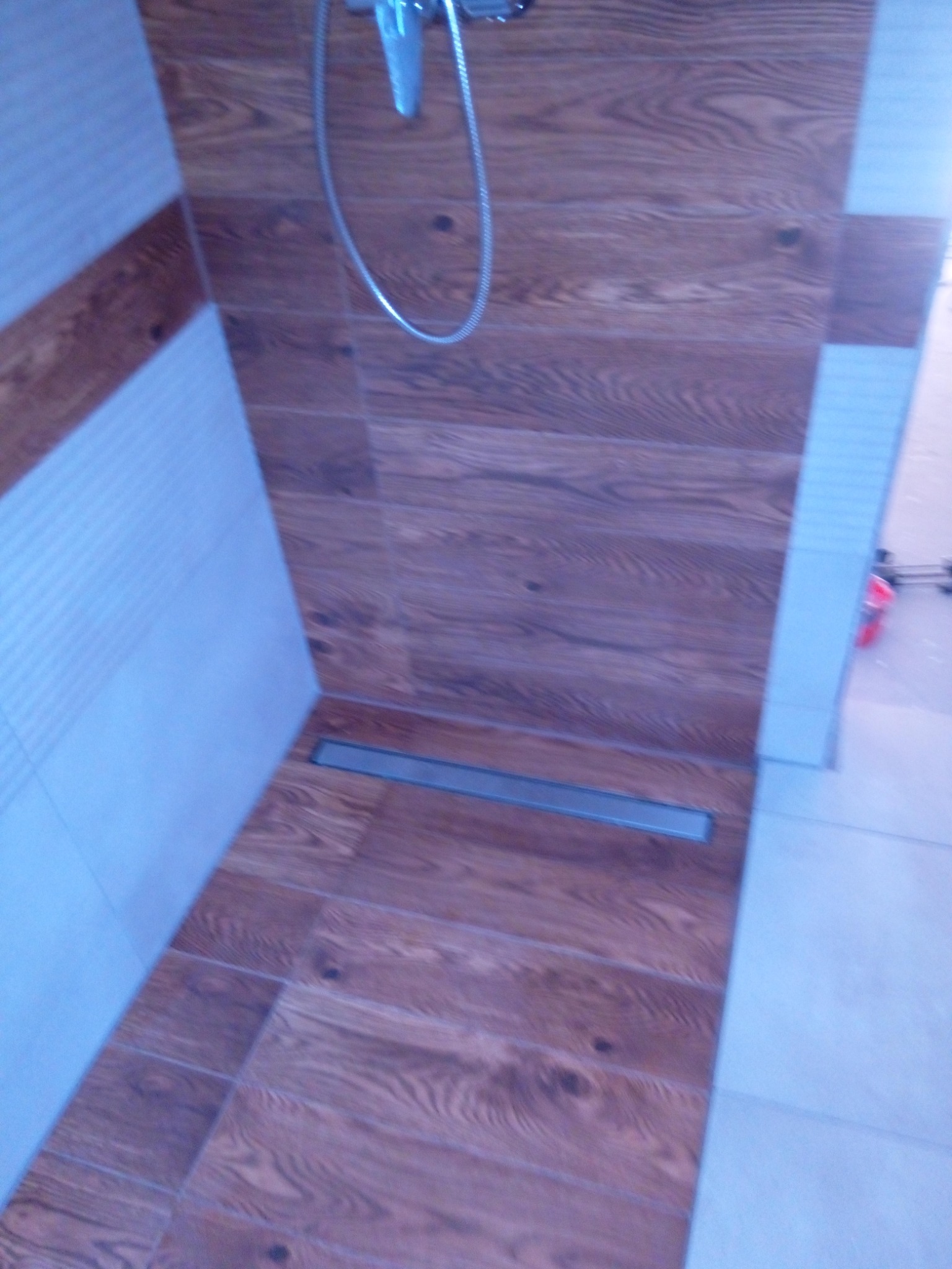 Prysznic z odpływem liniowym, wykończony płytkami imitującymi drewno na ścianach i podłodze, z chromowaną słuchawką prysznicową.