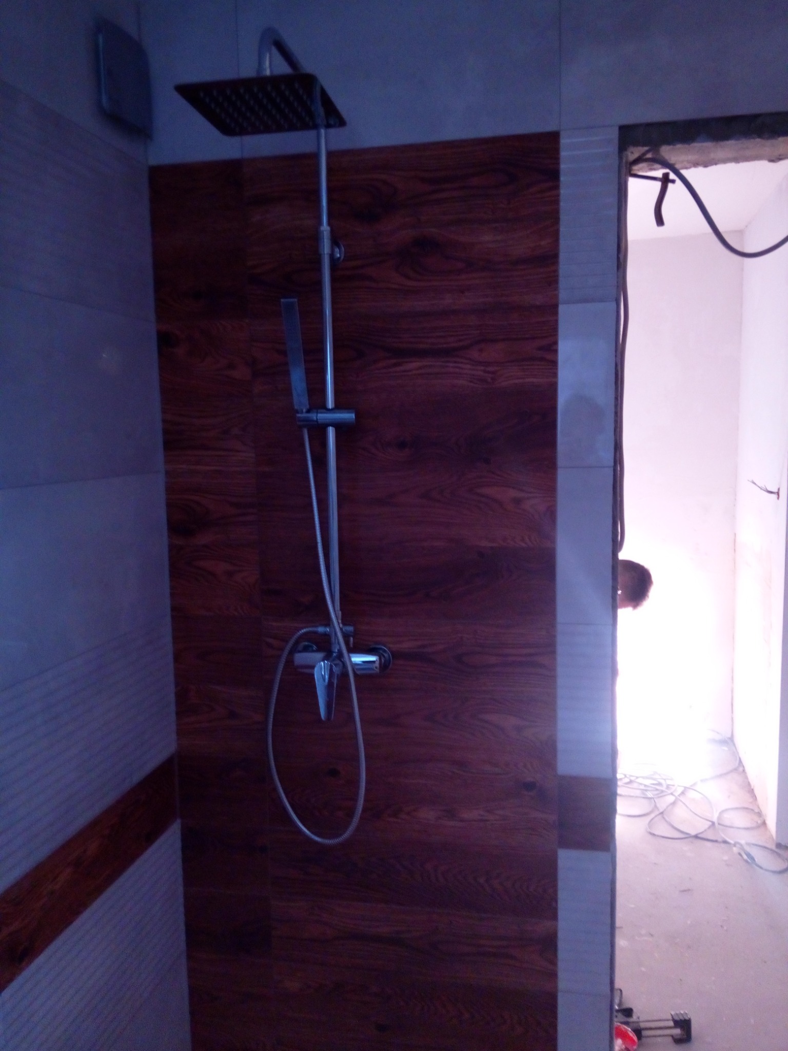 Kabina prysznicowa z panelem imitującym drewno, chromowana bateria z deszczownicą, widoczne przewody elektryczne i fragment osoby w przejściu do innego pomieszczenia.