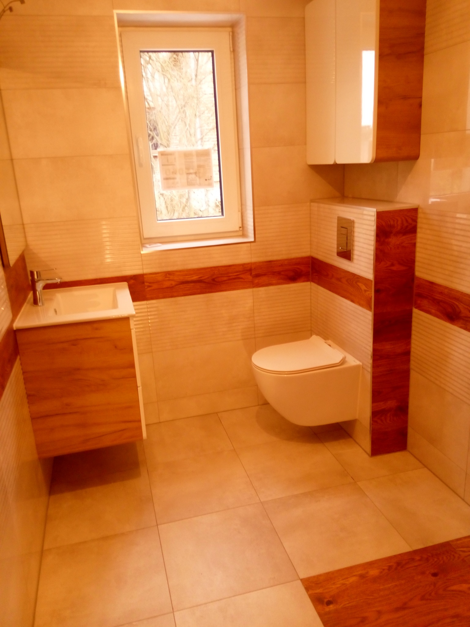 Jasna łazienka z drewnianymi akcentami: umywalka z szafką, podwieszana toaleta, szafka wisząca, beżowe płytki z poziomym pasem drewna, okno z widokiem na drzewa.