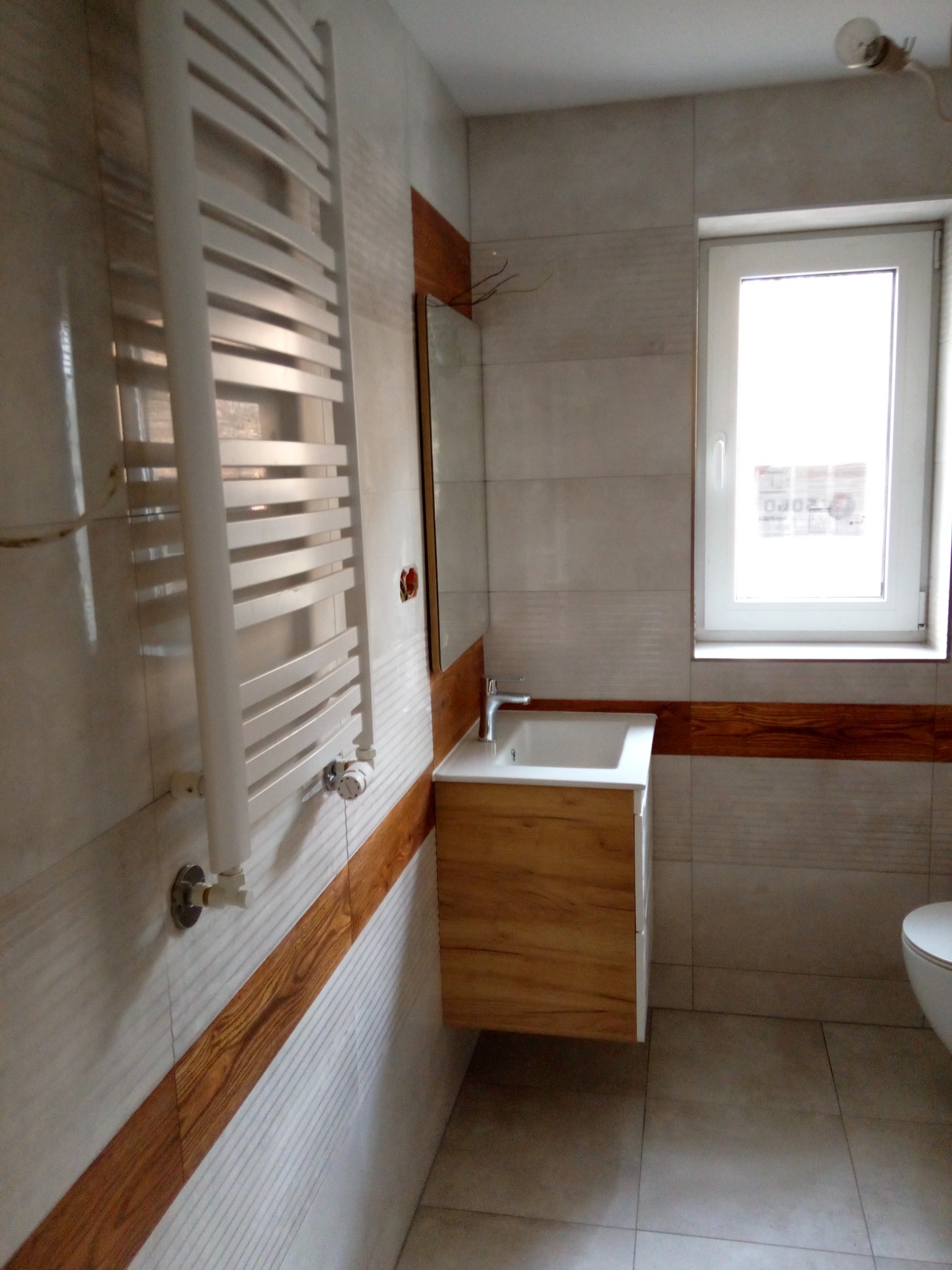 Nowoczesna łazienka z wiszącą szafką pod umywalką z frontami imitującymi drewno, grzejnikiem drabinkowym i białymi płytkami z poziomym pasem dekoracyjnym w kolorze drewna.